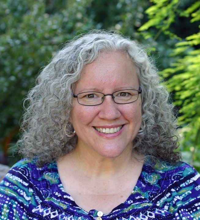 Woman with gray curly hair and glasses smiles outdoors.