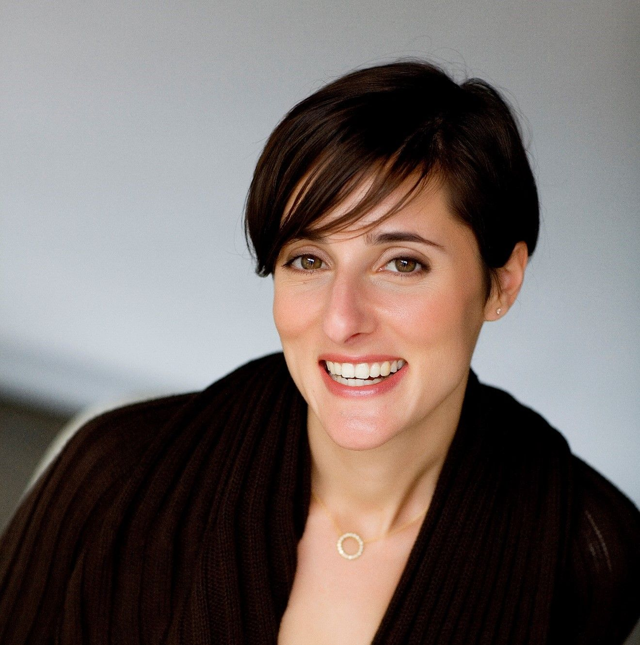 Woman with short dark hair, smiling, wearing a brown sweater and a gold necklace.