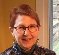Woman with short red hair and glasses smiles. Wearing a patterned jacket and olive turtleneck. Indoor setting.