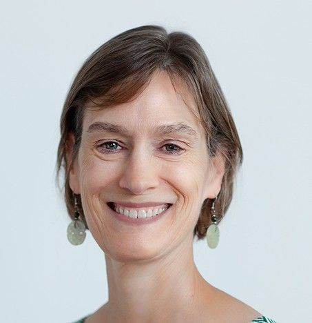 Woman with curly blonde hair and glasses smiles. Wearing green earrings and a white shirt.