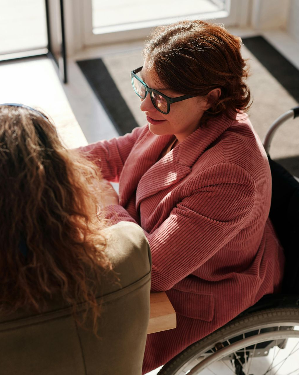 Woman in wheelchair with glasses, talking to another person at a table, indoors.