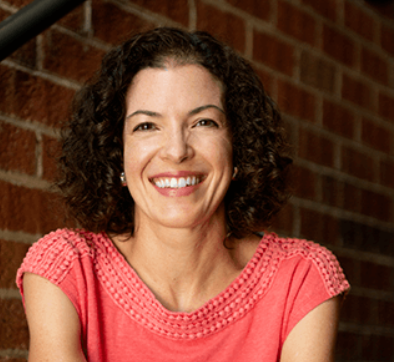 Woman with curly hair smiles, seated on steps against brick wall; wears coral top, arms crossed.