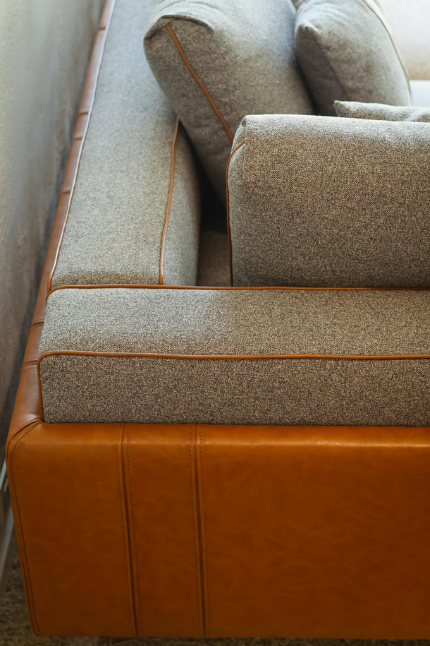 Close-up of a gray upholstered sofa with orange leather base and piping, against a white wall.