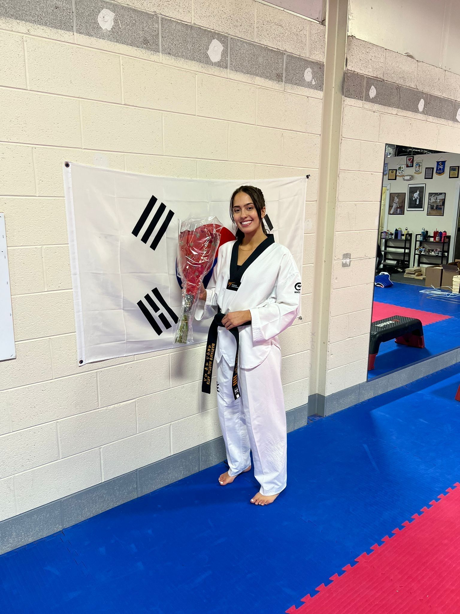 A woman in a taekwondo uniform is holding a bouquet of flowers in front of a flag.