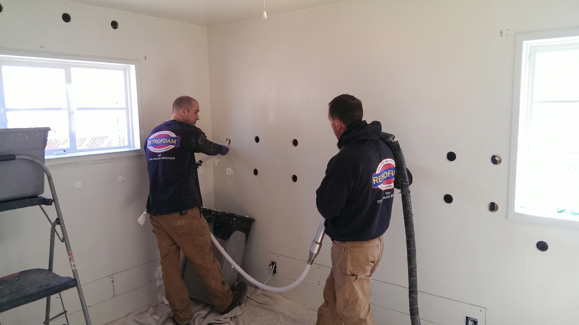 Two workers in hoodies inspecting a white room wall with tools and cables near windows.