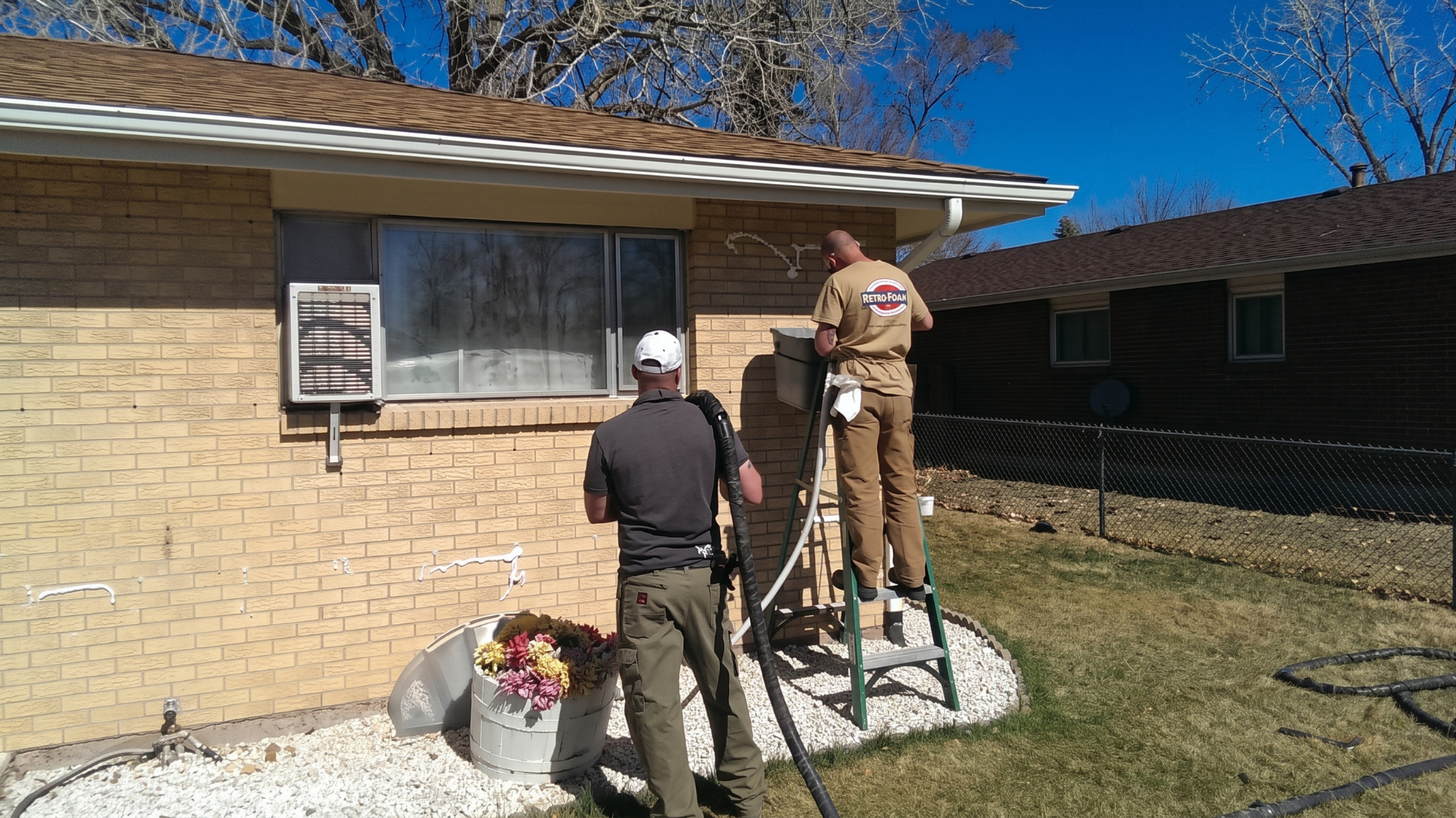 Two workers installing holiday lights on a brick house by a window, one on a ladder