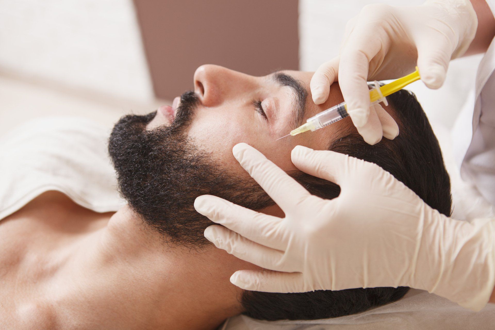 A man receives a facial injection from gloved hands. He lies down with eyes closed. The setting is a medical office.