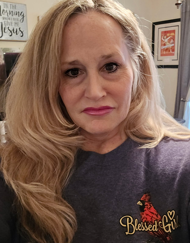Woman with long blonde hair, wearing a grey shirt, looking at the camera. In the background is a framed picture and a sign that reads 