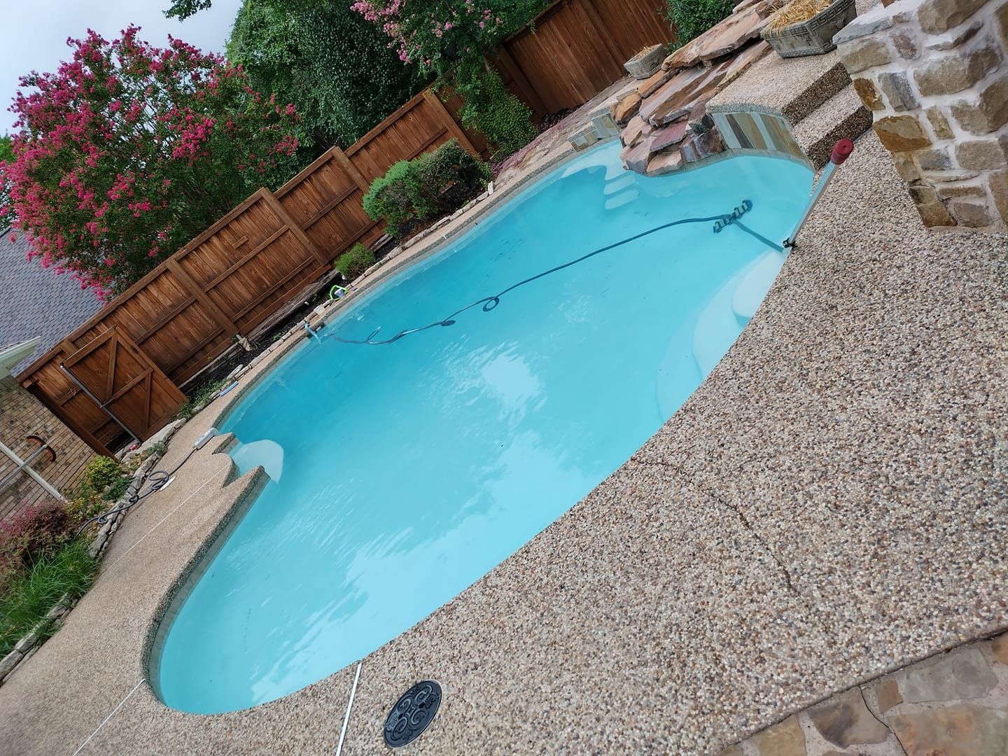 Backyard swimming pool with turquoise water, stone deck, and wooden fence under flowering trees