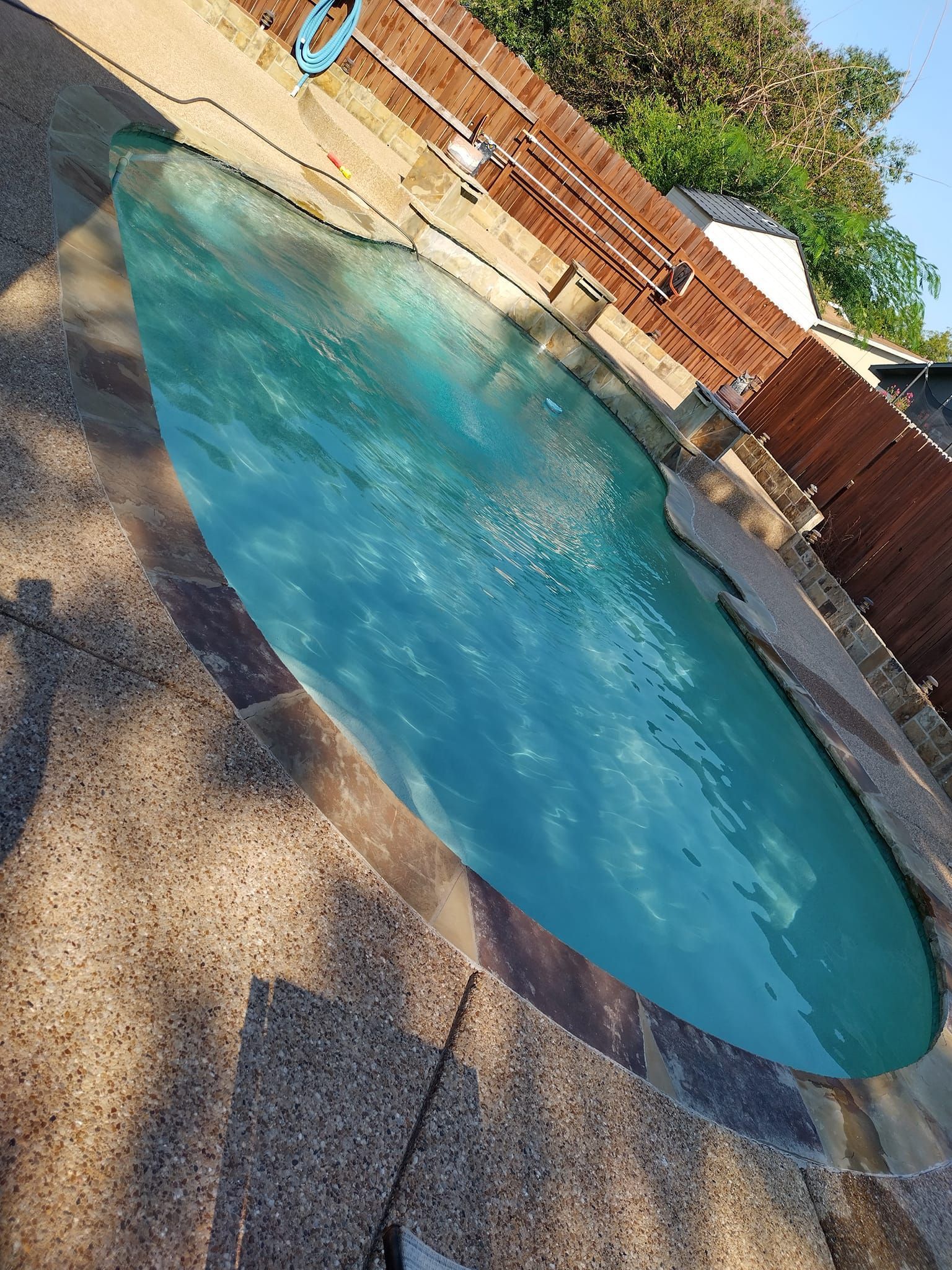 Backyard swimming pool with blue water, brick patio, and wooden fence under sunlight