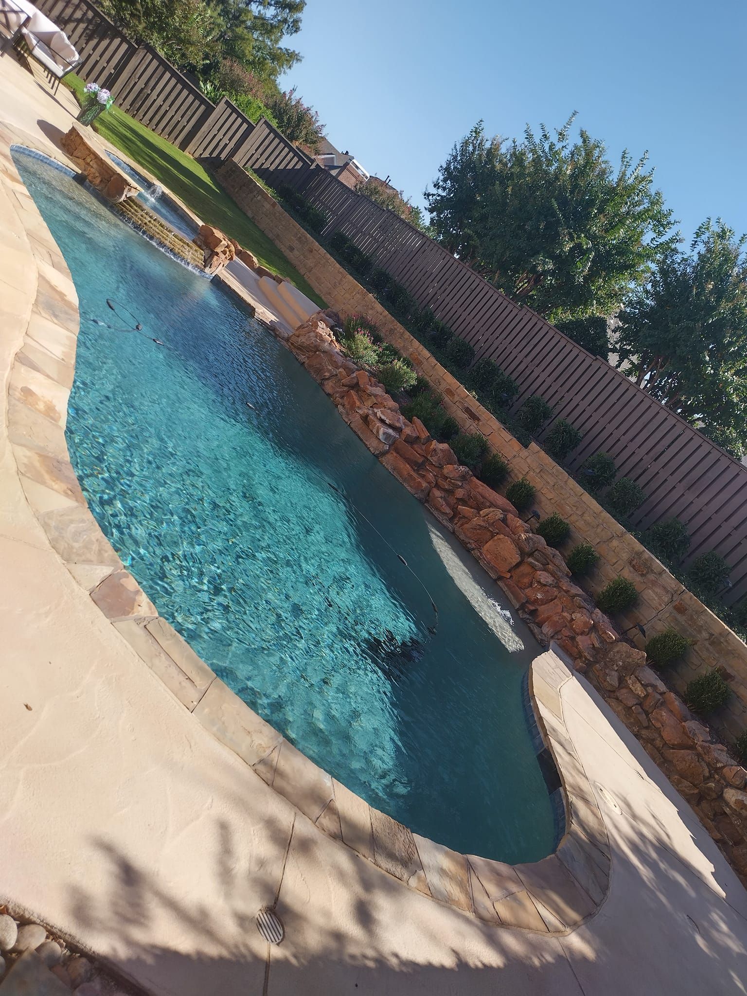 Backyard swimming pool with blue water, stone deck, brick wall, and trees in sunlight