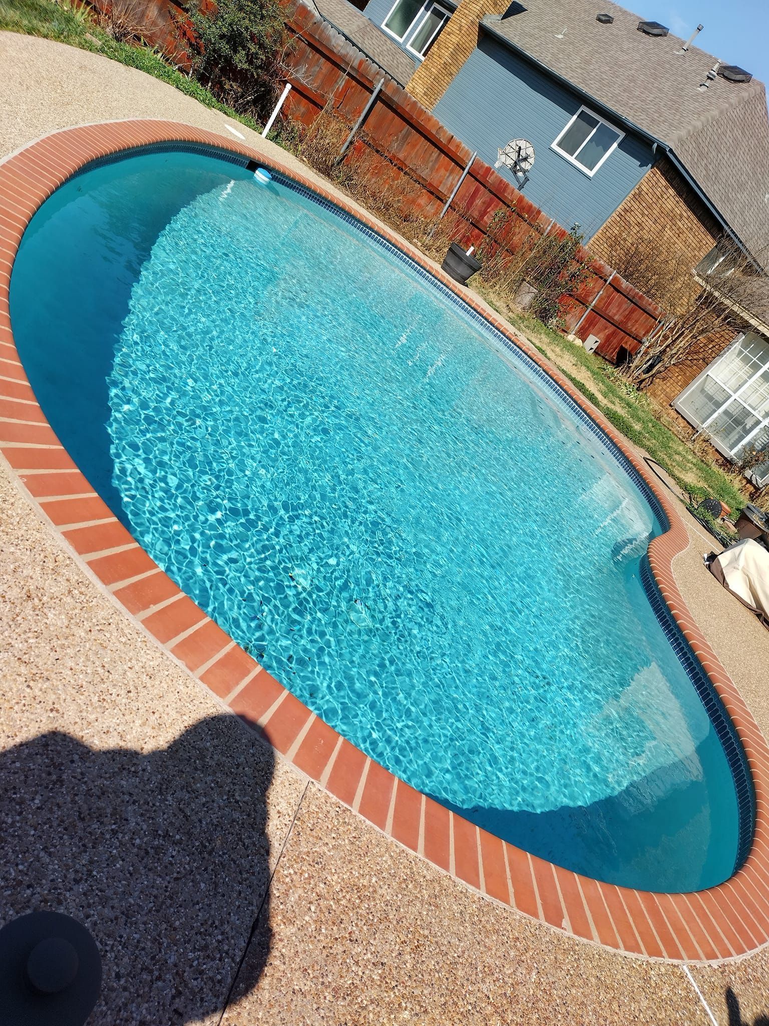 Backyard oval swimming pool with blue water, brick coping, and gravel patio beside a blue house