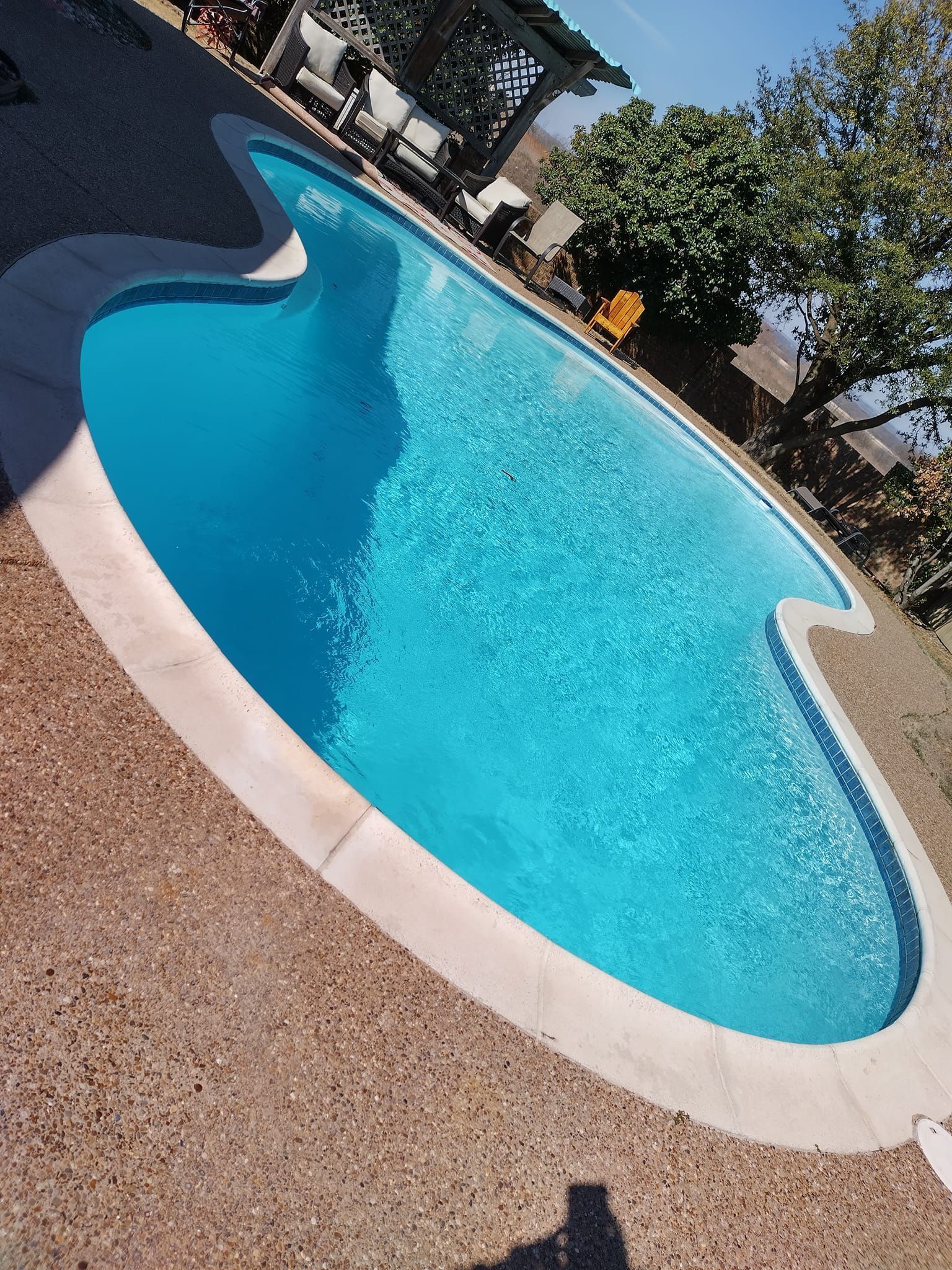 Blue swimming pool with white coping and a pebbled deck, viewed at an angle in bright sunlight