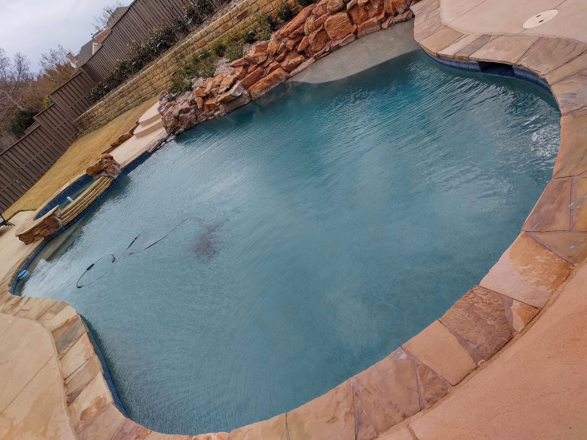 Backyard swimming pool with blue water, tan stone border, and a rock waterfall feature