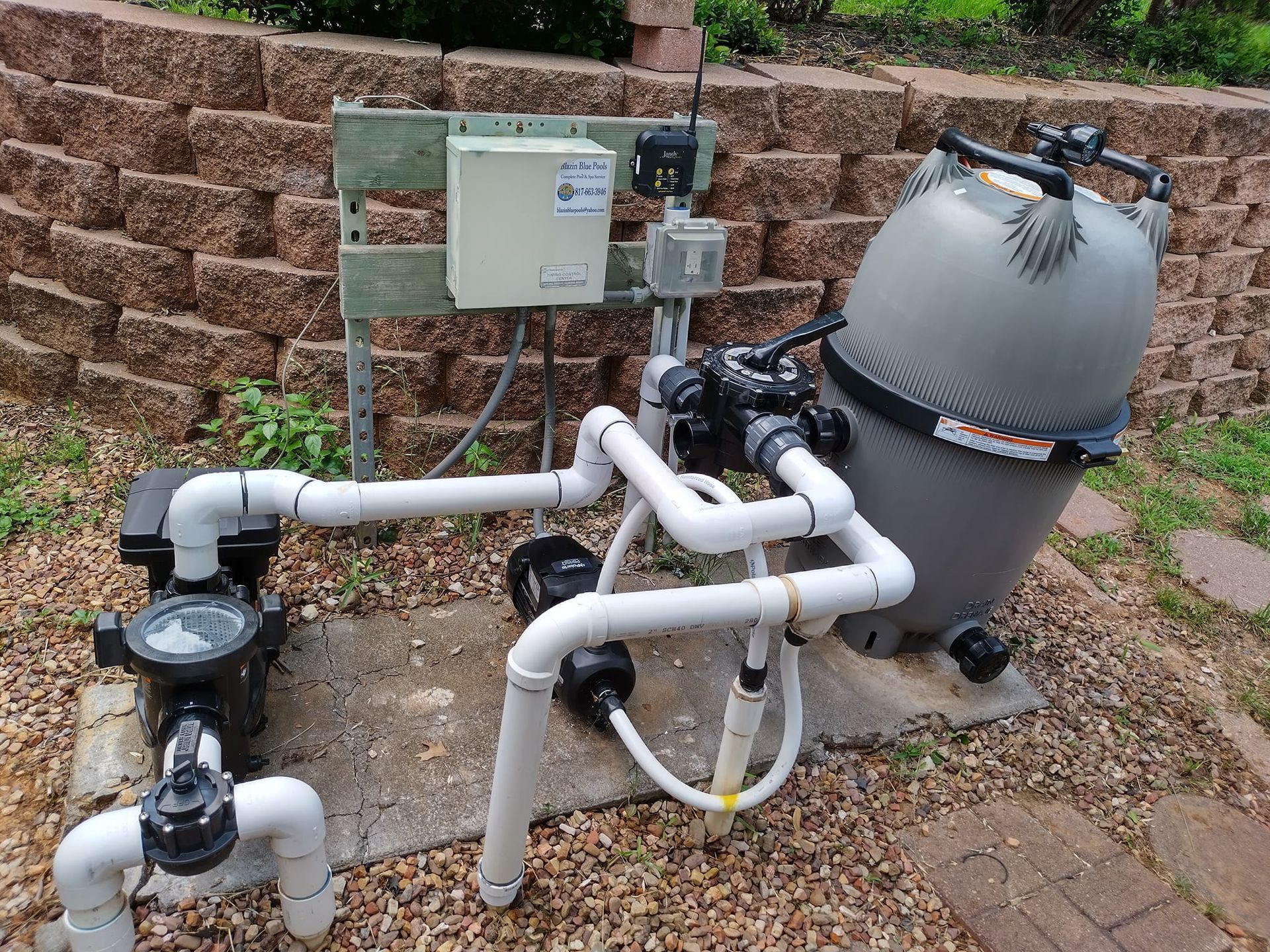Outdoor pool filtration and pump equipment with pipes, control box, and sand filter beside a stone wall.