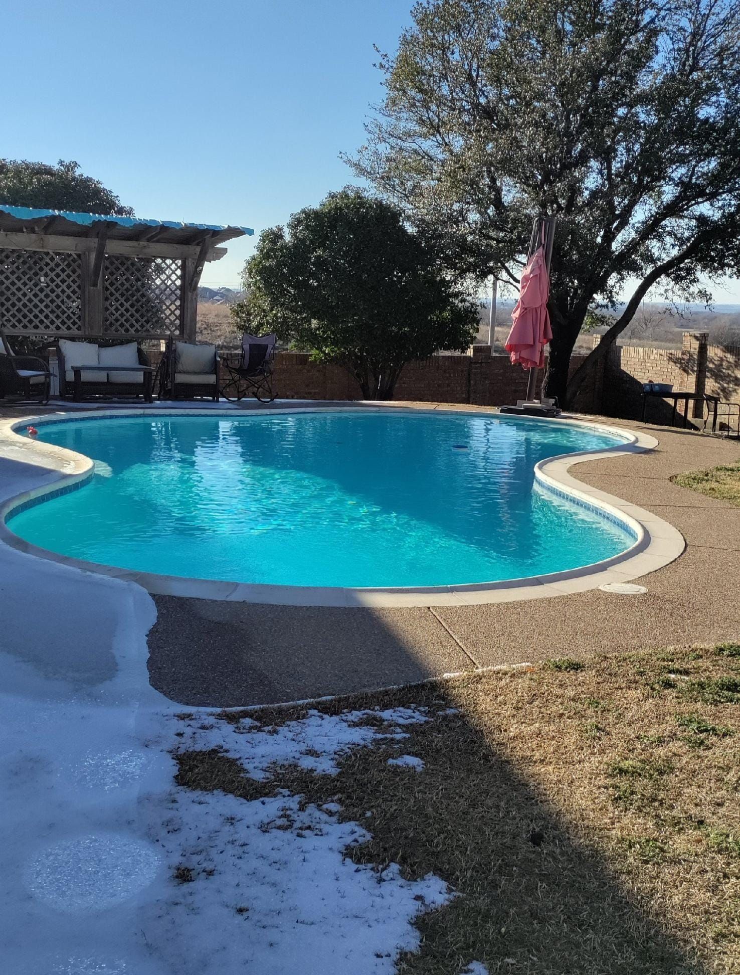 Backyard swimming pool with turquoise water, patio seating, and a pink umbrella under trees on a sunny day