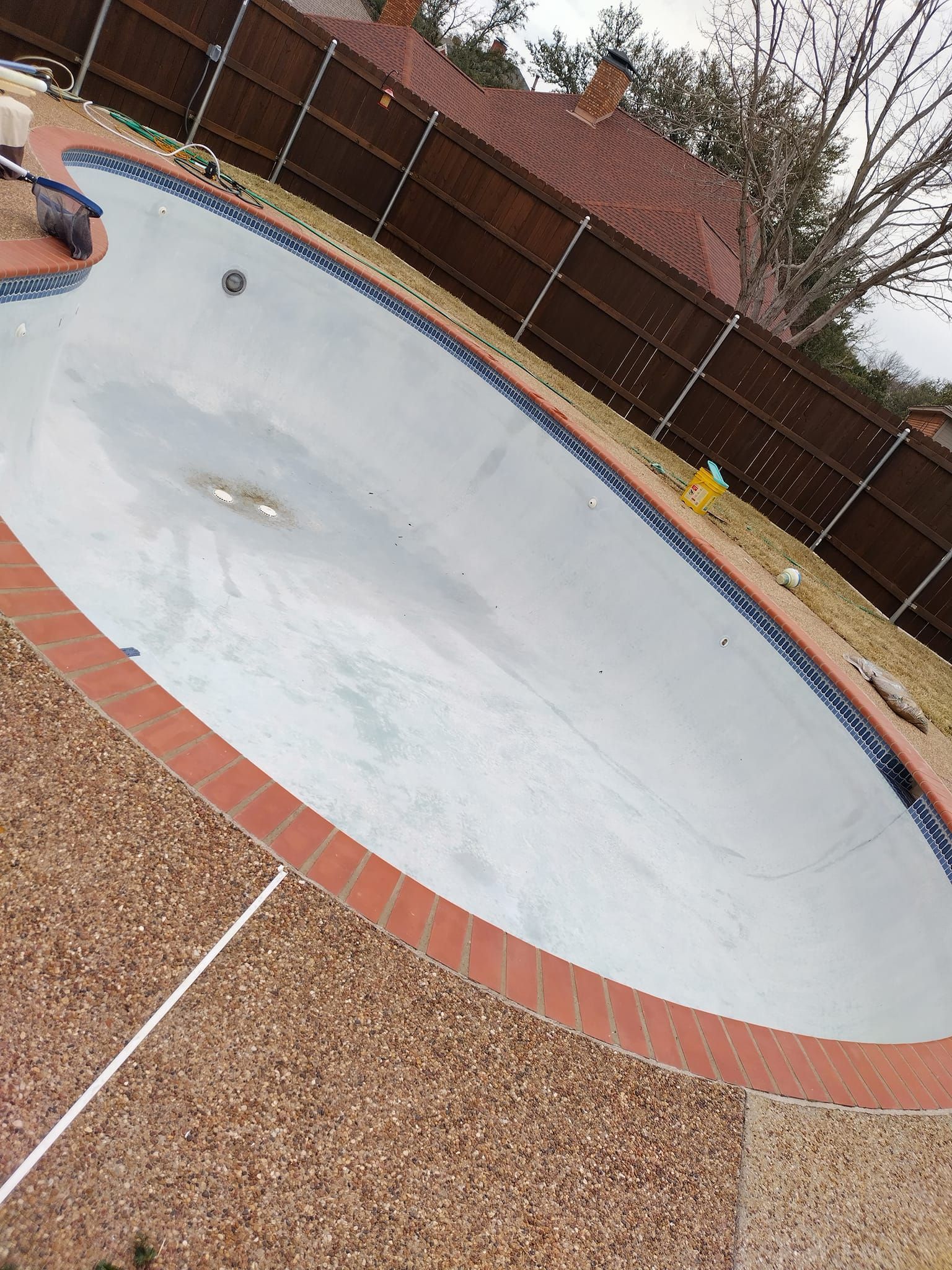 Empty drained swimming pool with faded white surface, brick coping, and brown fence in a backyard.