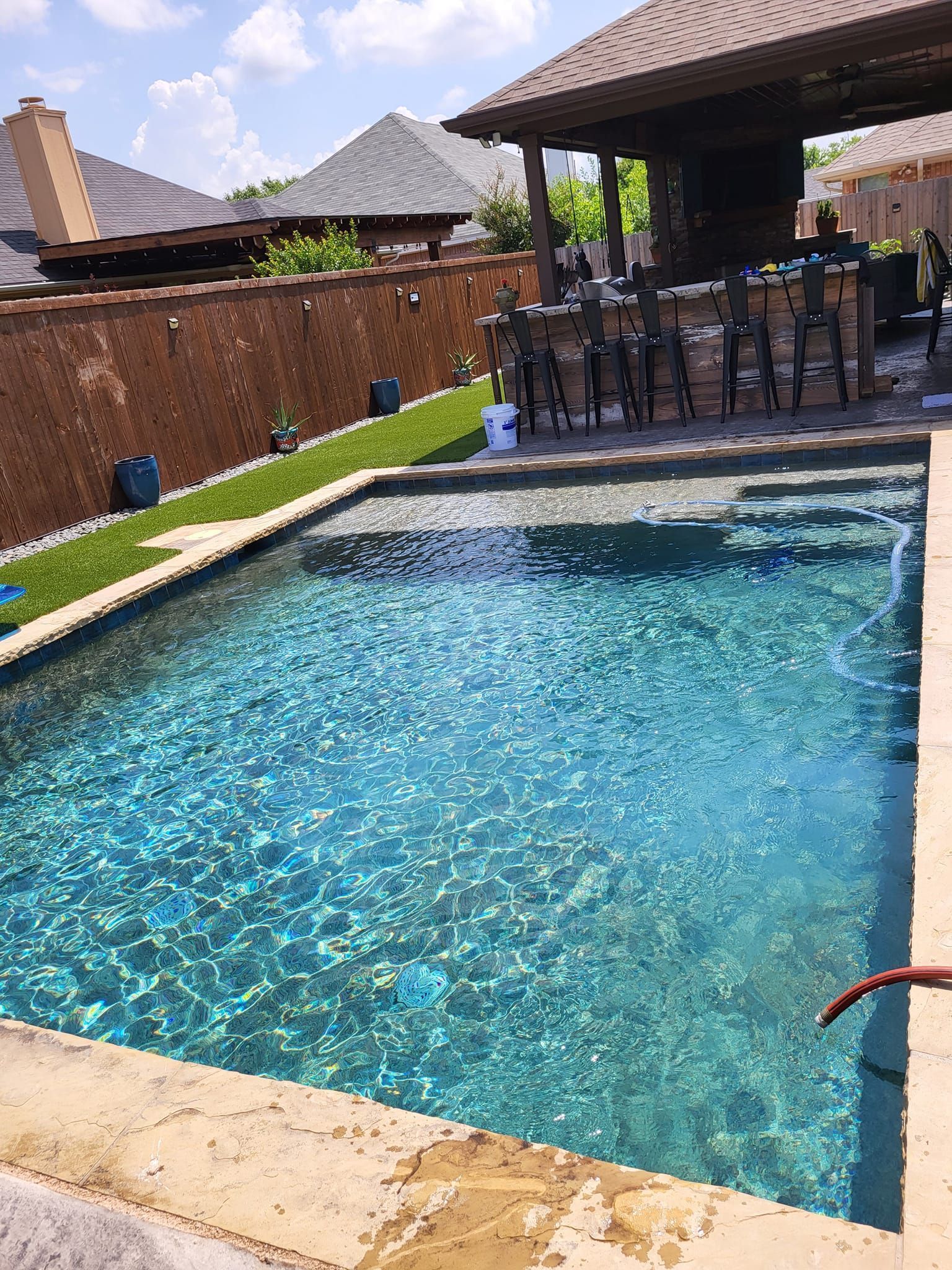 Blue backyard swimming pool beside a patio with outdoor dining furniture and a brick privacy wall