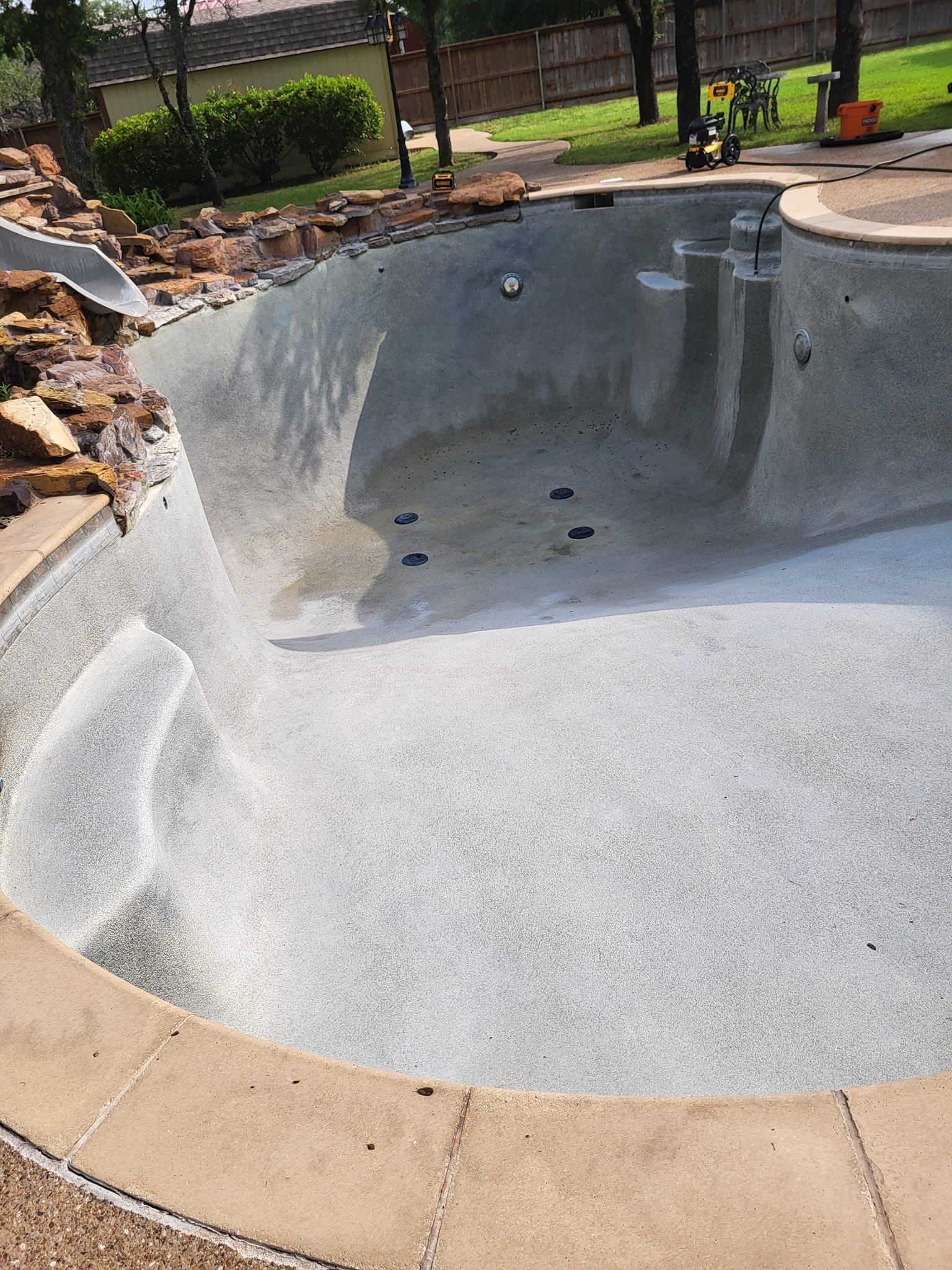 Empty drained swimming pool with a light gray bottom and tan coping beside a landscaped yard