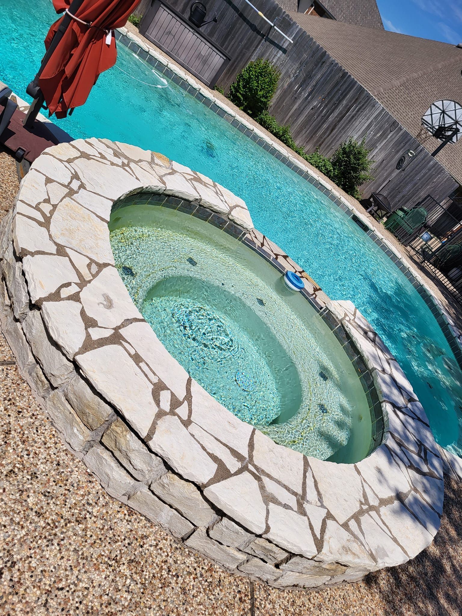 Backyard hot tub beside a turquoise pool, with gravel and a wooden fence in the background.