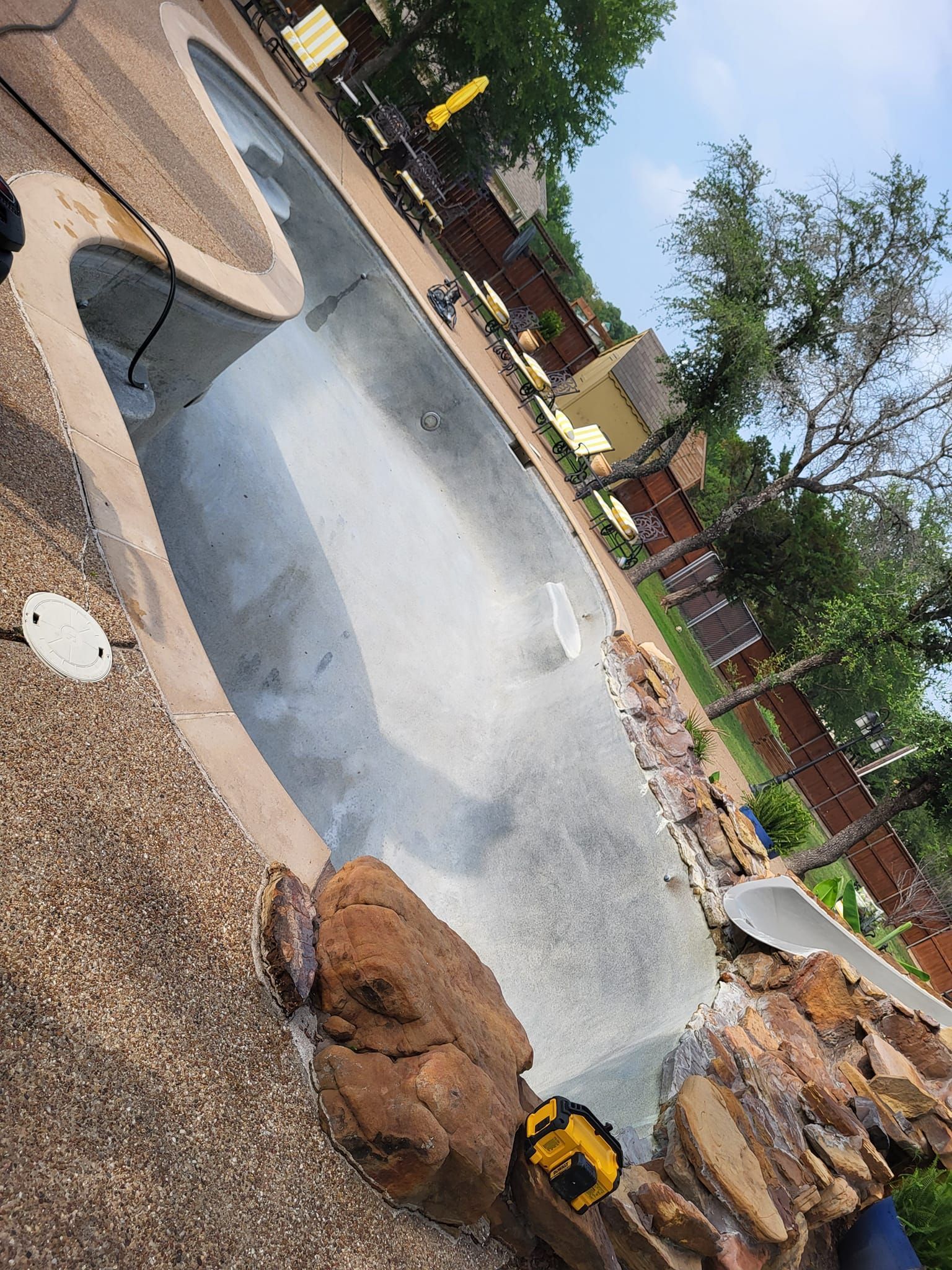 Backyard pool under construction, with concrete shell, rocks, and a yellow excavator nearby