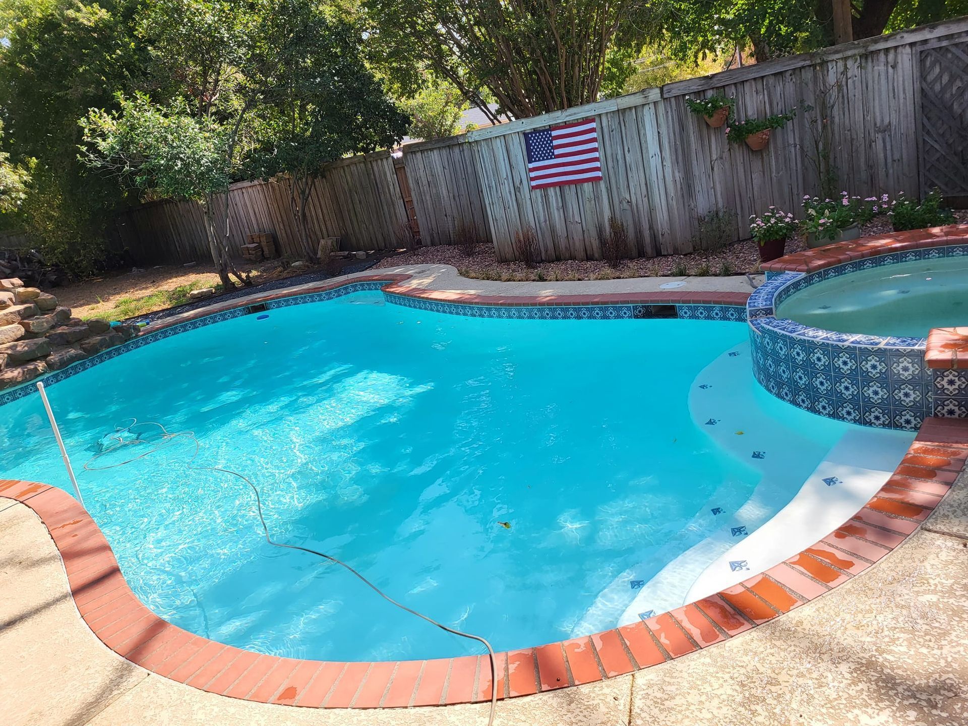 Curved backyard swimming pool with turquoise water, brick coping, and a wooden fence with an American flag.