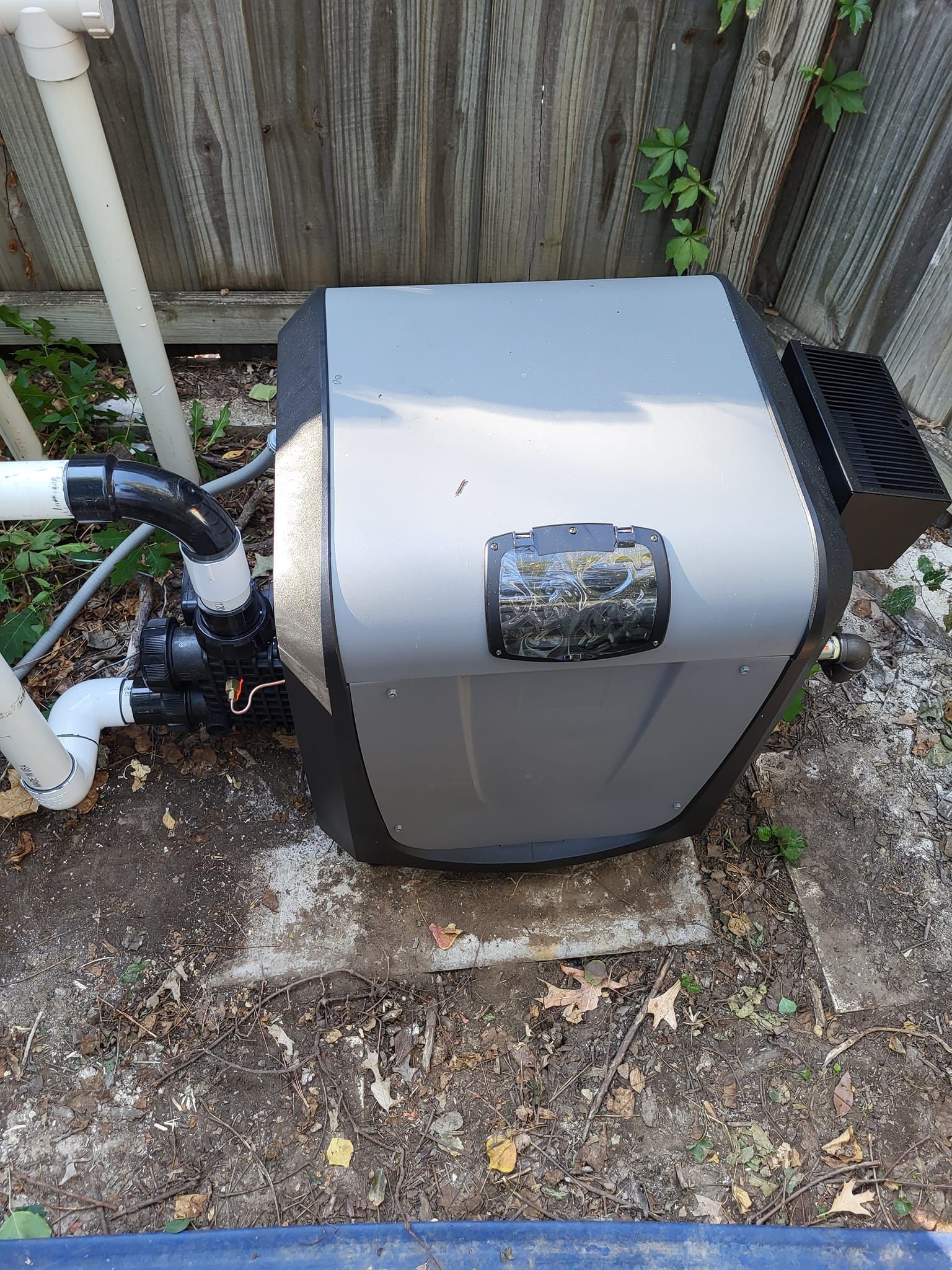 Gray outdoor utility box with pipes beside a wooden fence on a dirt patch