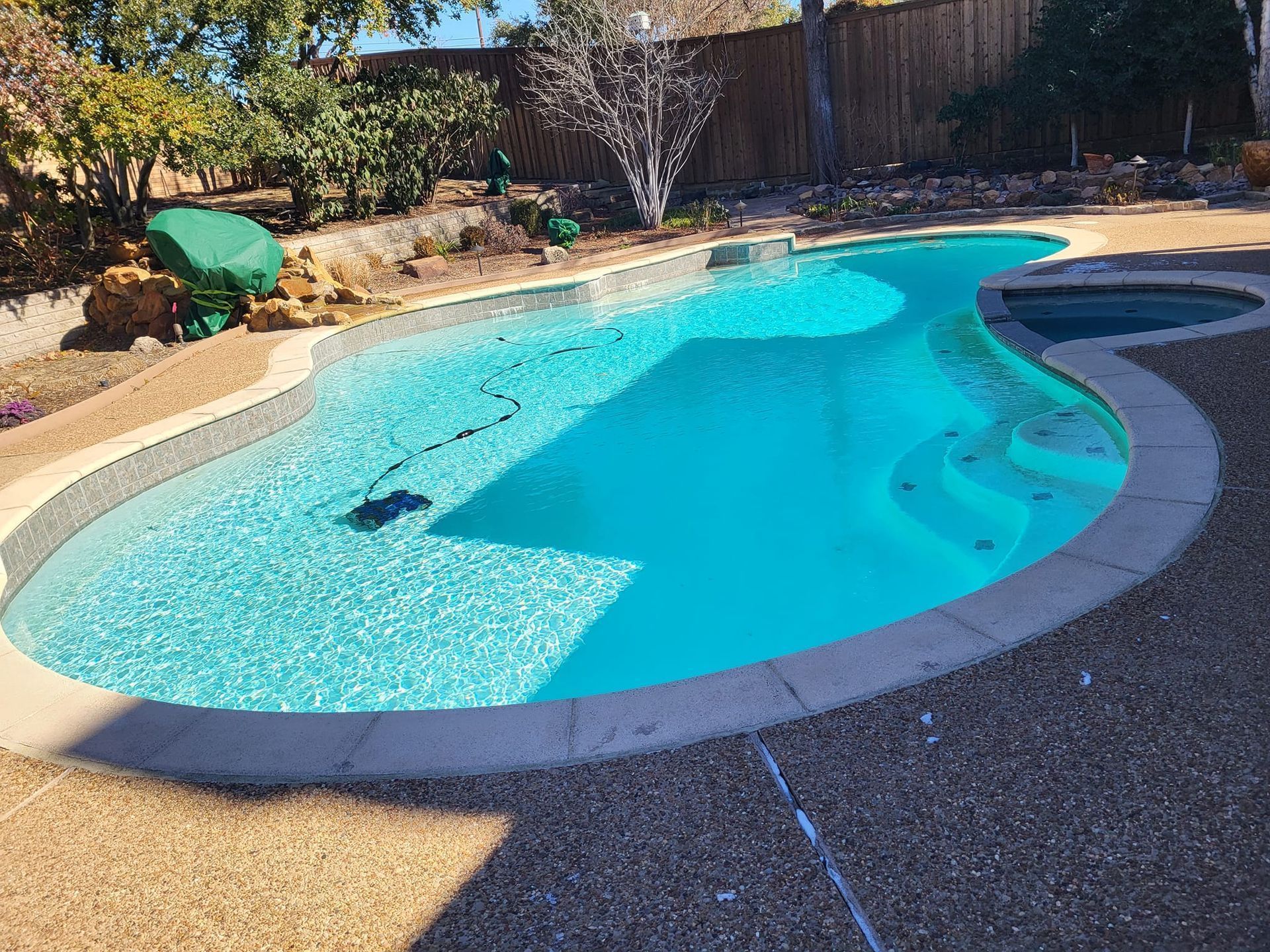 Curved backyard swimming pool with bright blue water, stone coping, and a shaded patio area.