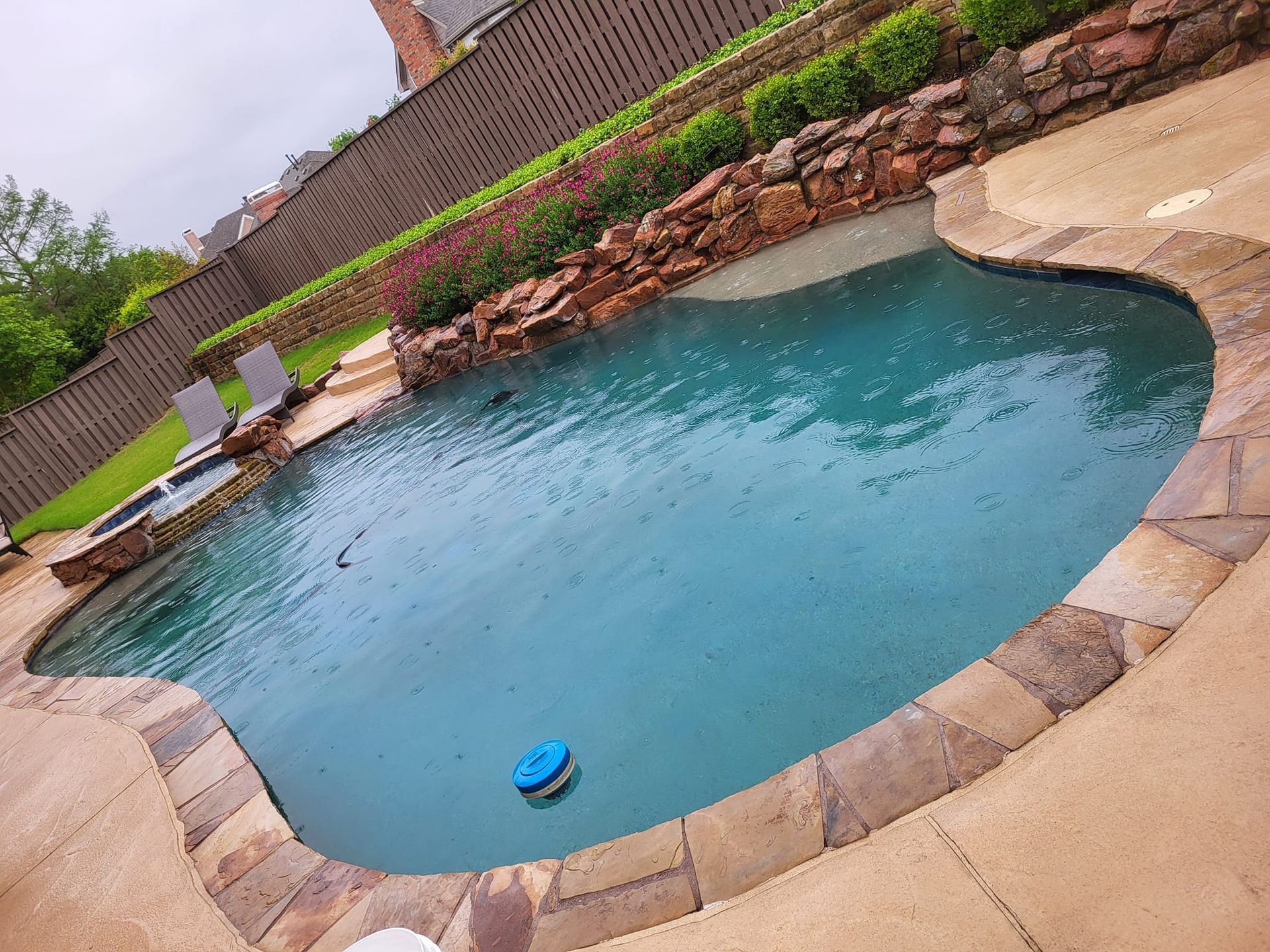 Backyard swimming pool with curved stone deck and brick wall, viewed at a slight angle