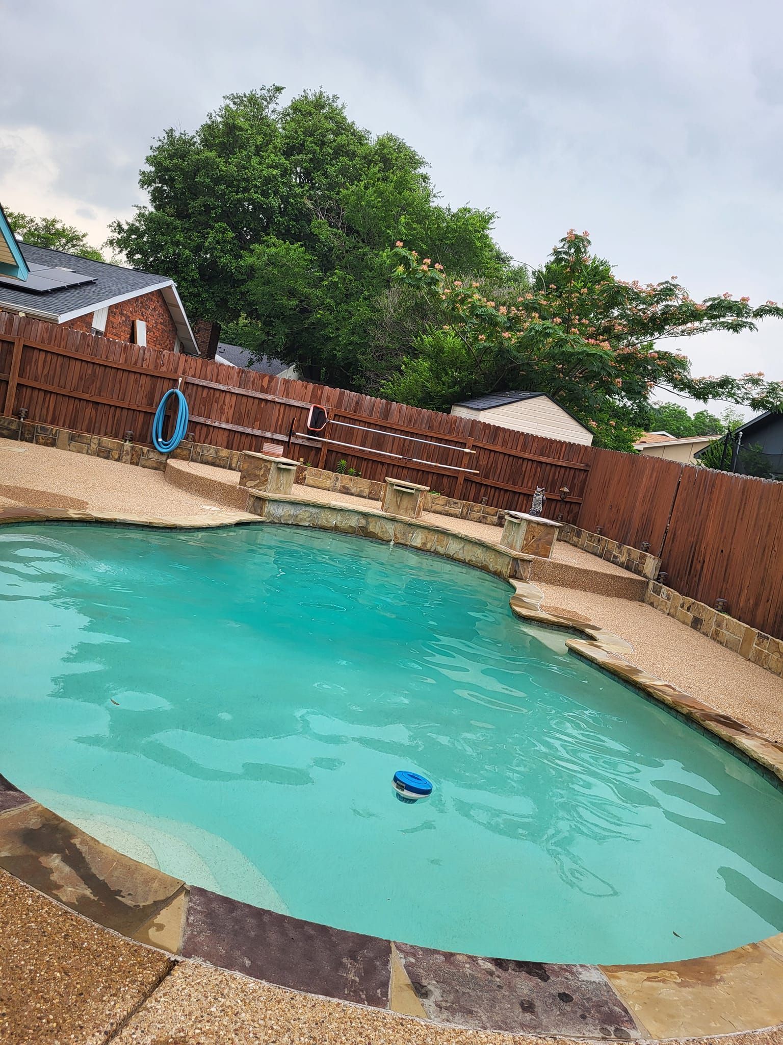 Backyard swimming pool with turquoise water, brick wall, and trees under a cloudy sky
