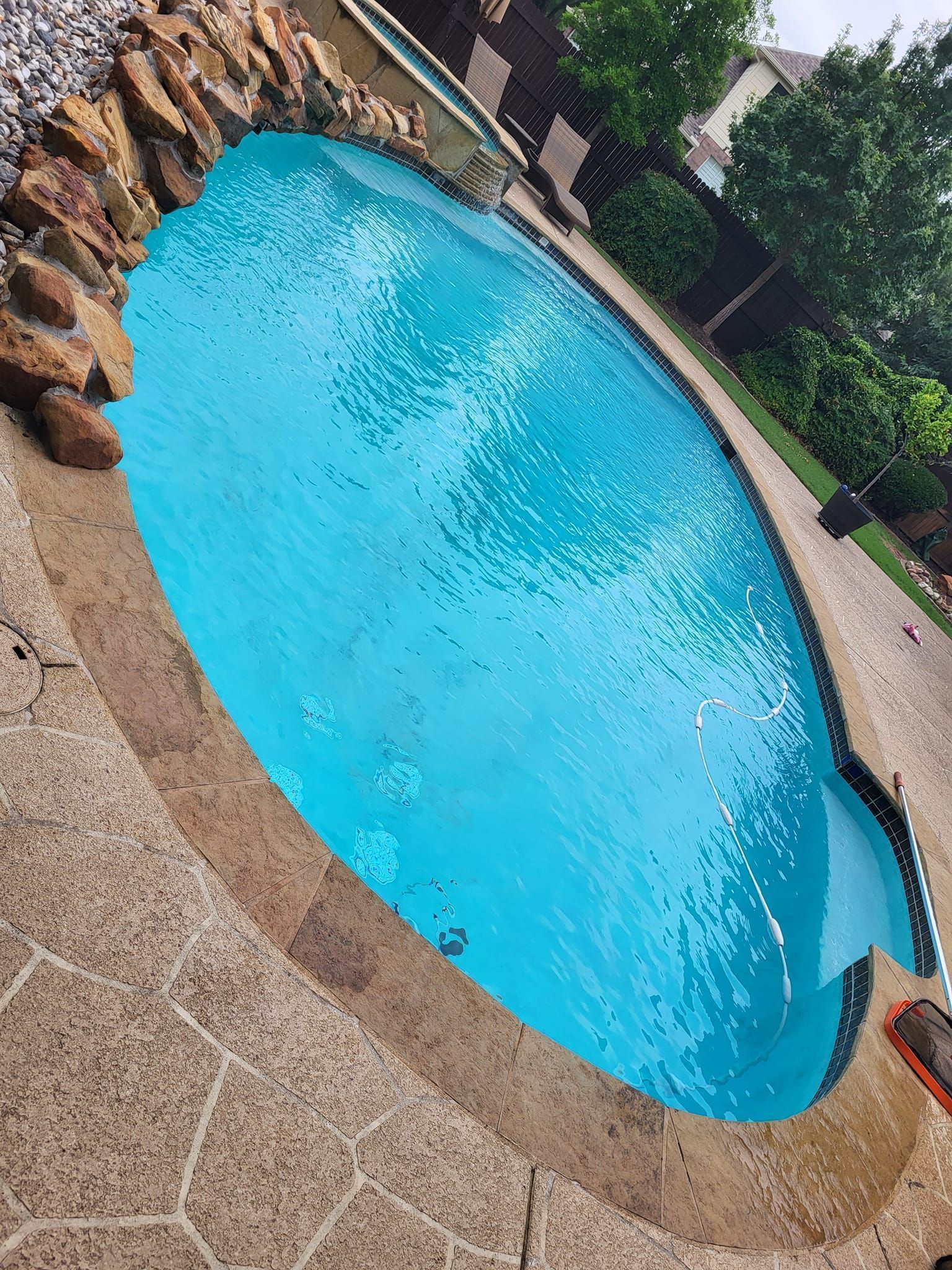 Blue swimming pool with stone deck and surrounding greenery, viewed from above at an angle