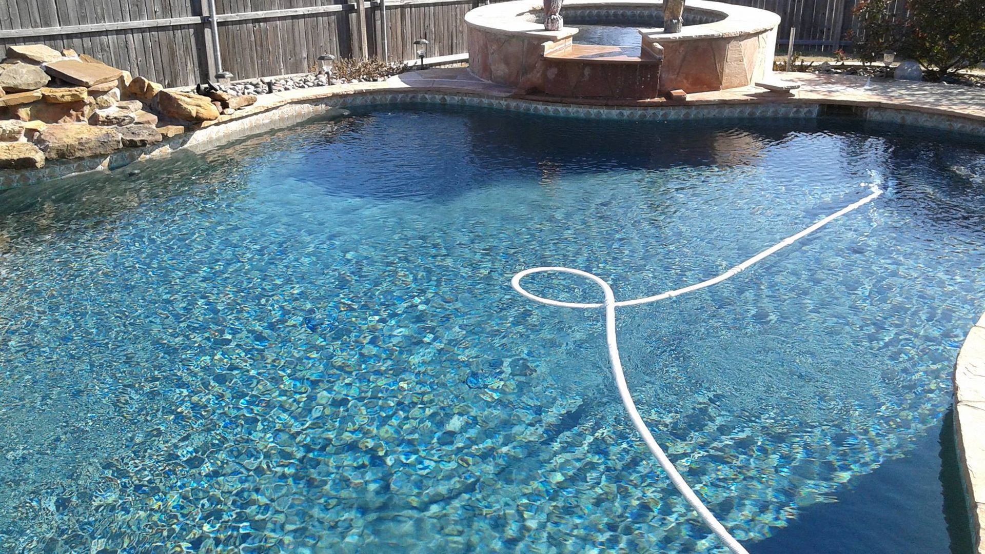 Backyard swimming pool with a white hose floating on the blue water, beside a stone hot tub and wooden fence
