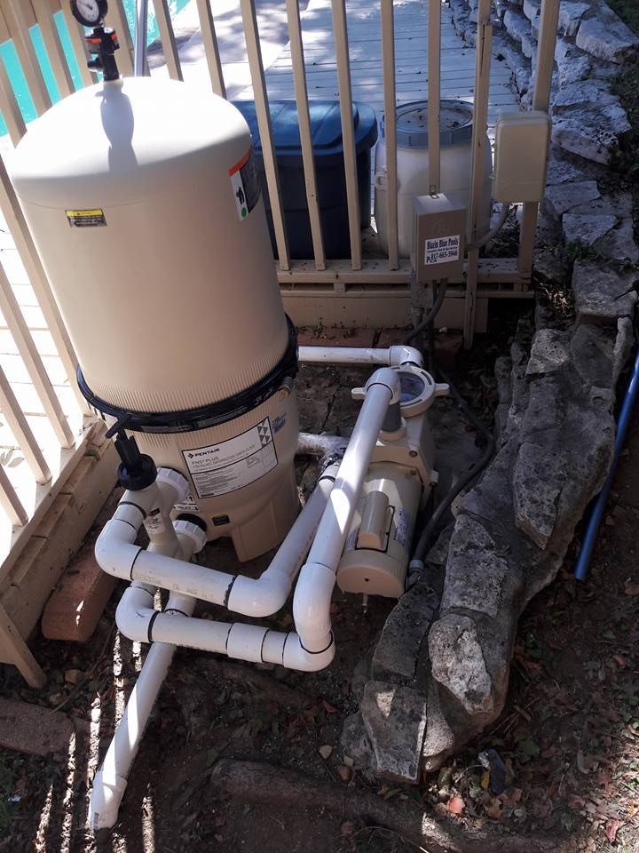 Outdoor pool pump and filter plumbing beside a fence and rocks.