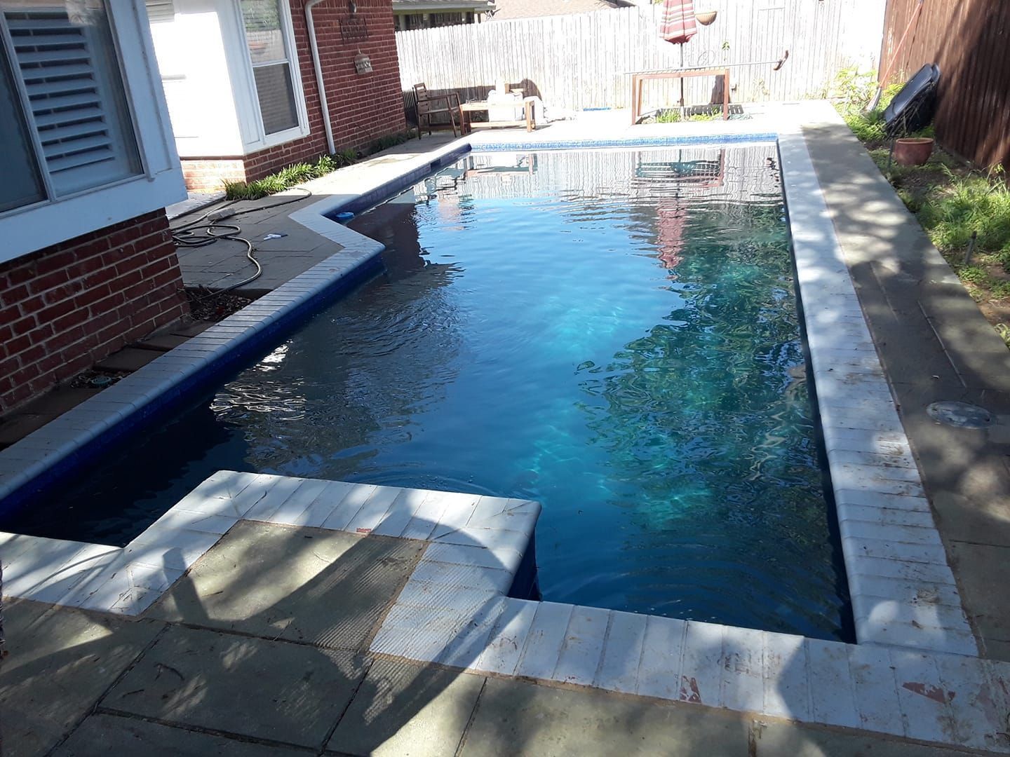 Narrow backyard swimming pool beside a brick house, with blue water and sunlit stone coping.
