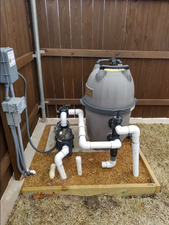 Outdoor pool filter and pump system beside a wooden fence, with white pipes and electrical boxes.