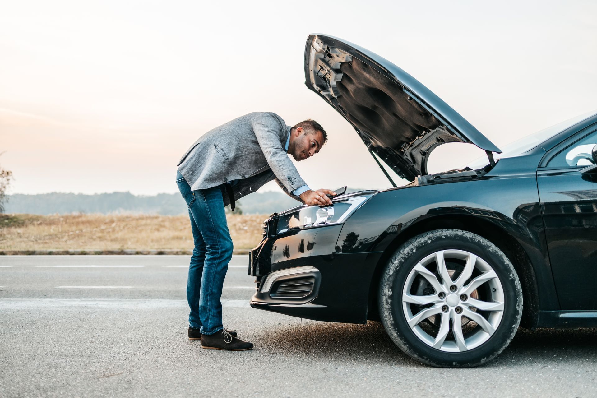 A person in business casual attire inspects the engine of a black car parked on the side of a road with the hood up.
