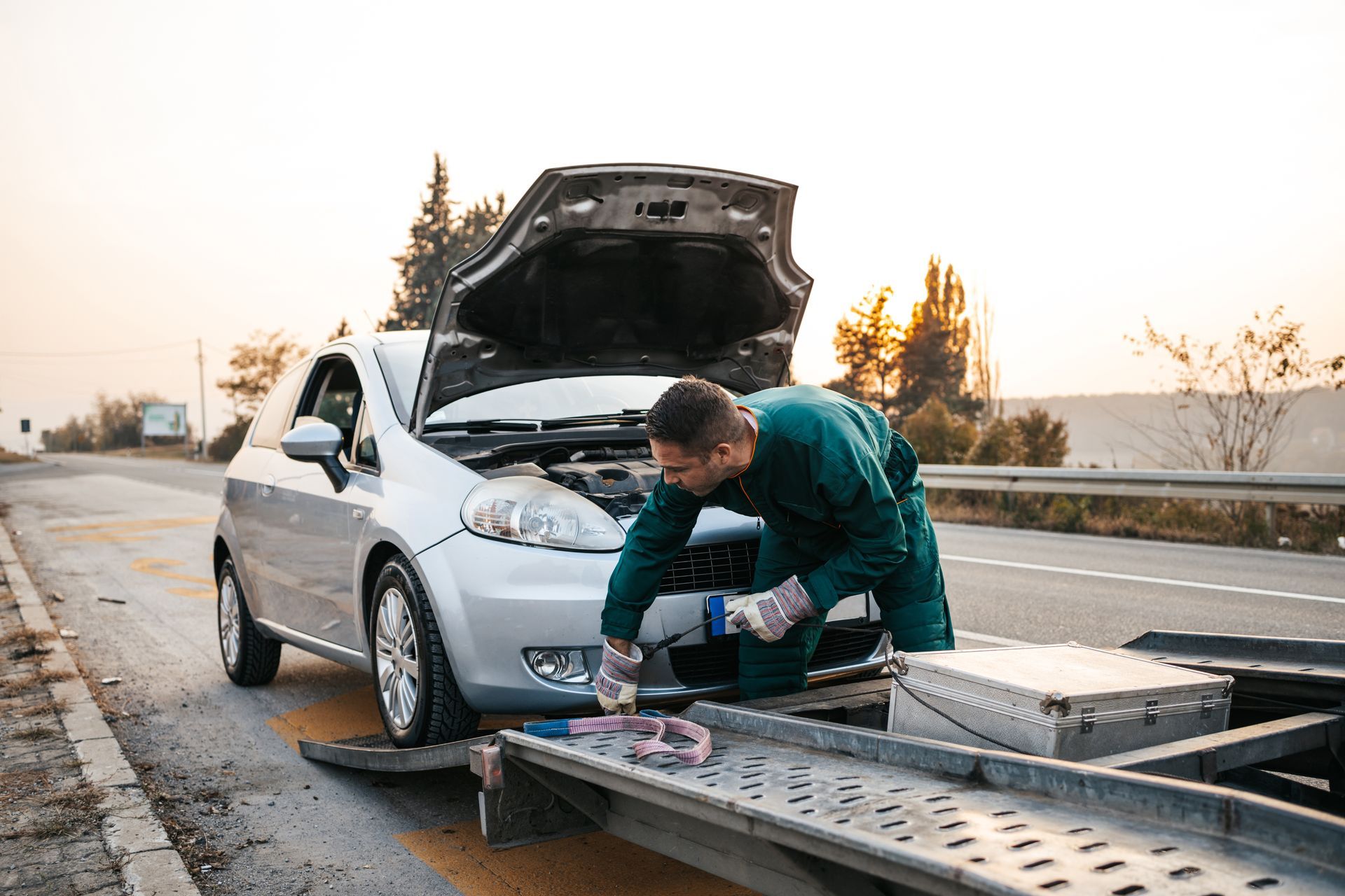 A worker in a green uniform attaches a strap to the front of a silver car being loaded onto a tow truck on the roadside.