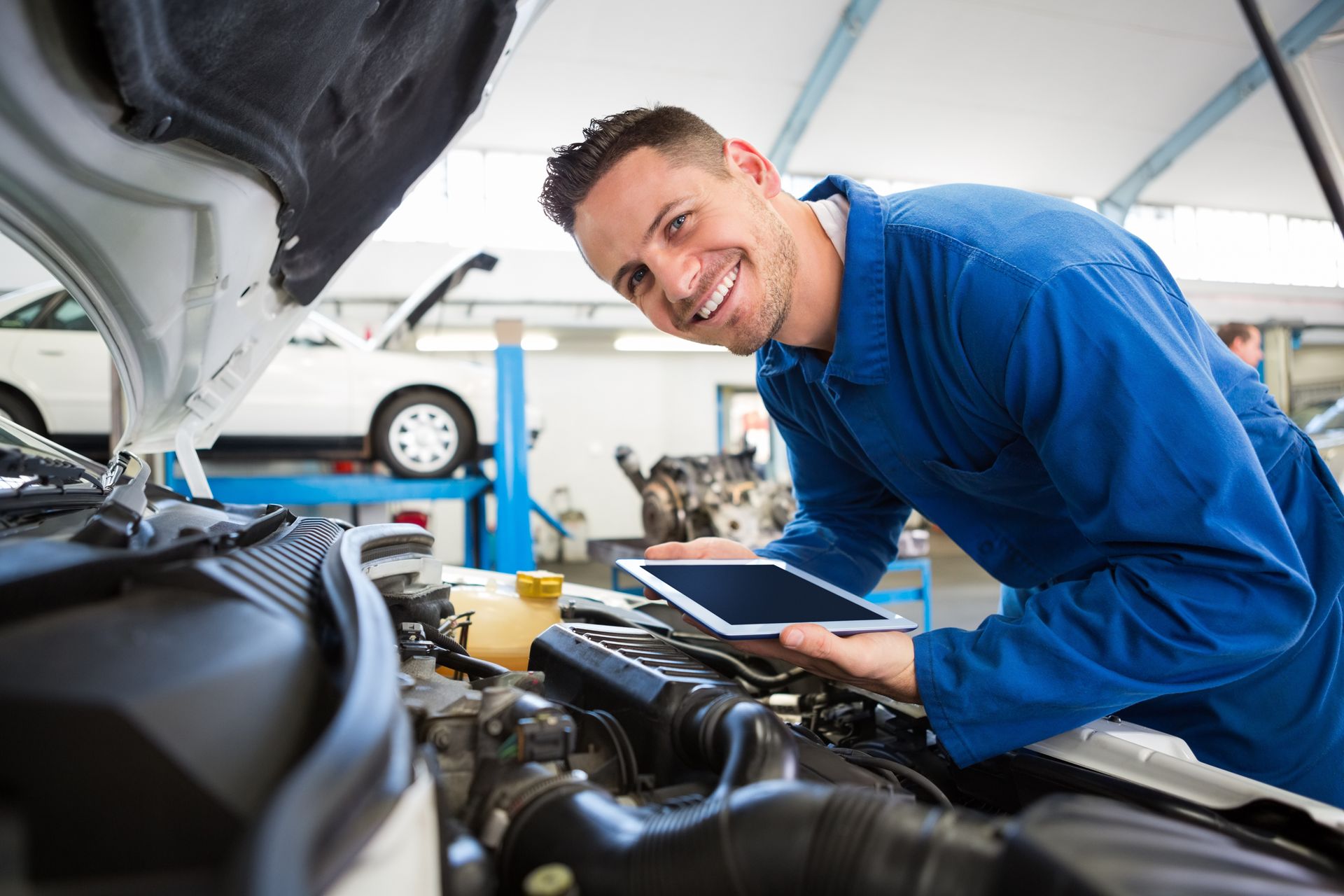 A smiling mechanic in a blue uniform holds a tablet while inspecting a car engine inside a repair shop. A smiling mechanic in a blue uniform holds a tablet while inspecting a car engine inside a repair shop.