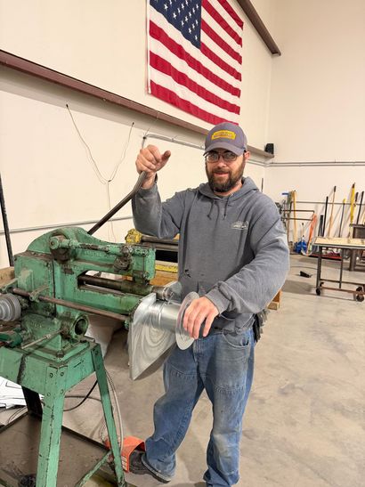 Man standing beside green metal machinery in a workshop, holding a metal part under a hanging American flag
