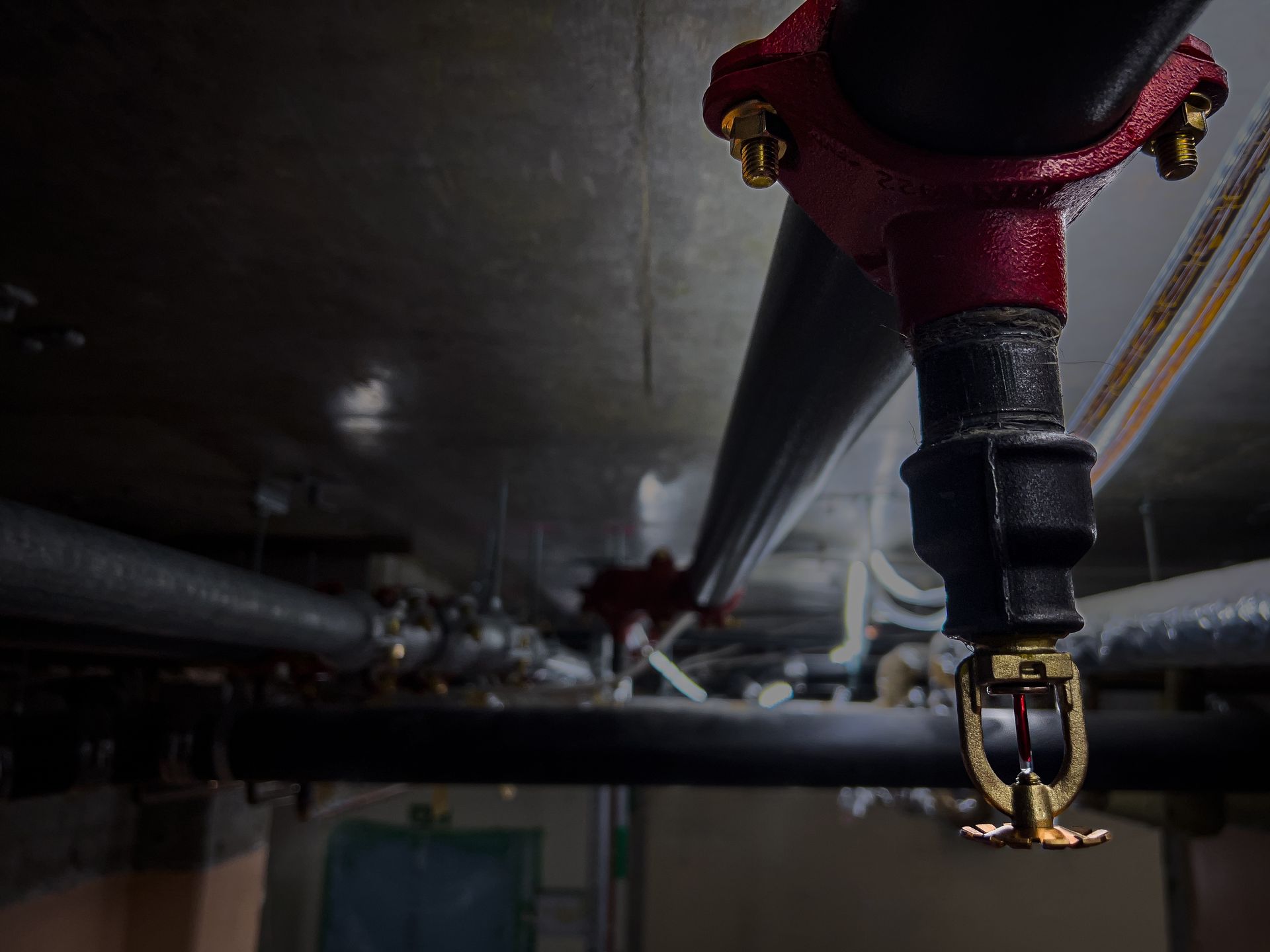 Close-up of a red industrial sprinkler pipe and valve assembly on a dark ceiling, with pipes receding into the distance