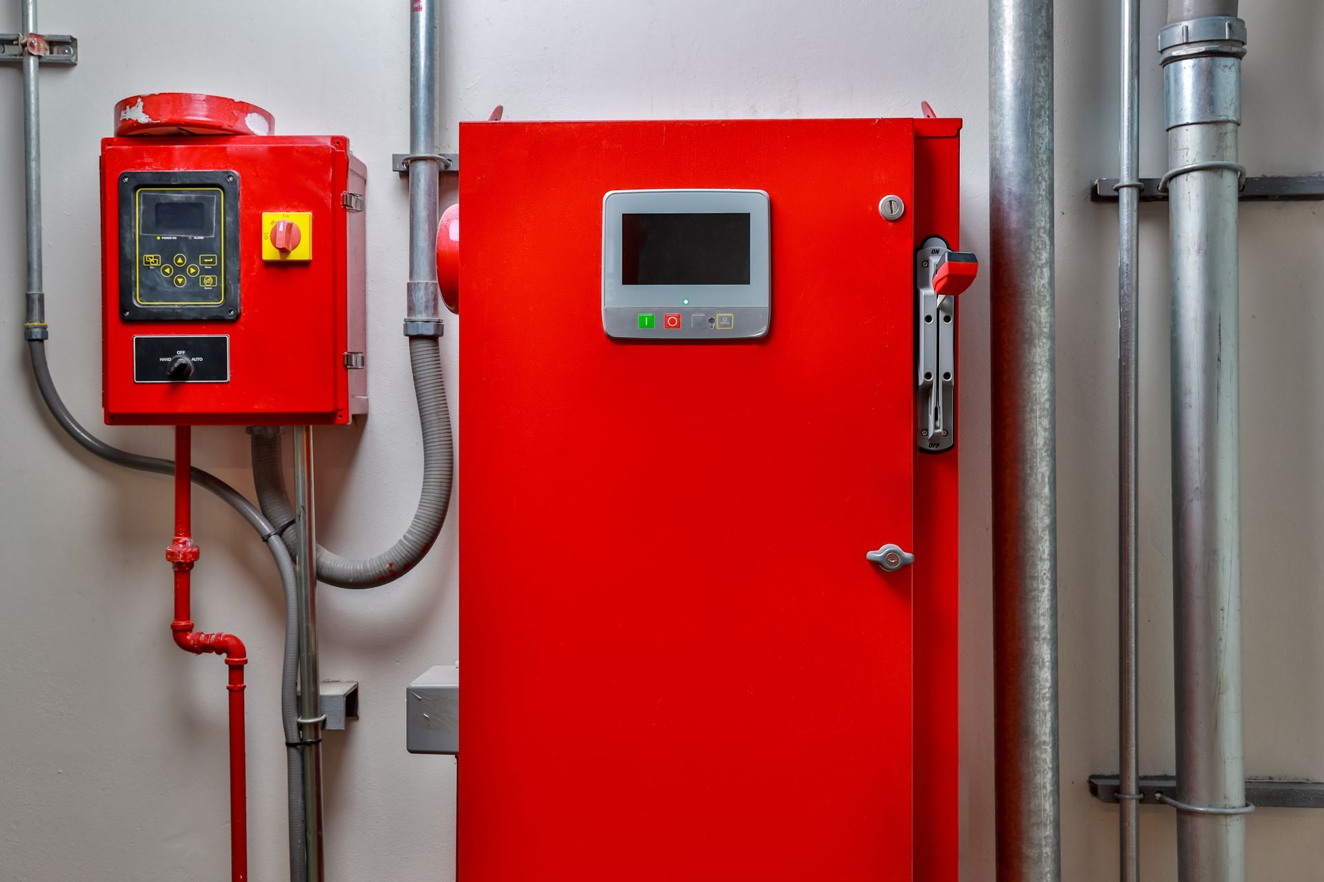Red industrial control panel and door-mounted display with pipes and conduit on a gray wall