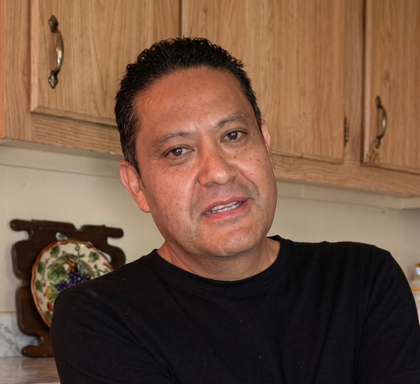 Man in black shirt standing in a kitchen, with wooden cabinets behind him.