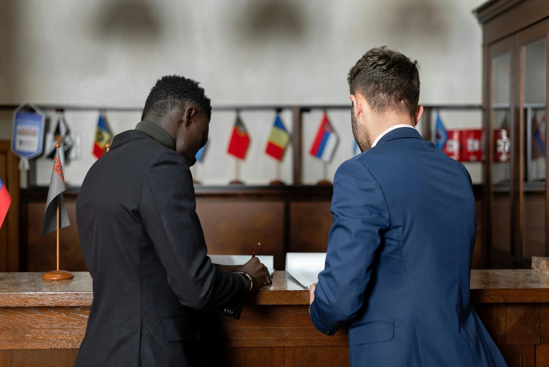 Two men in suits signing a document at a counter with small national flags in the background