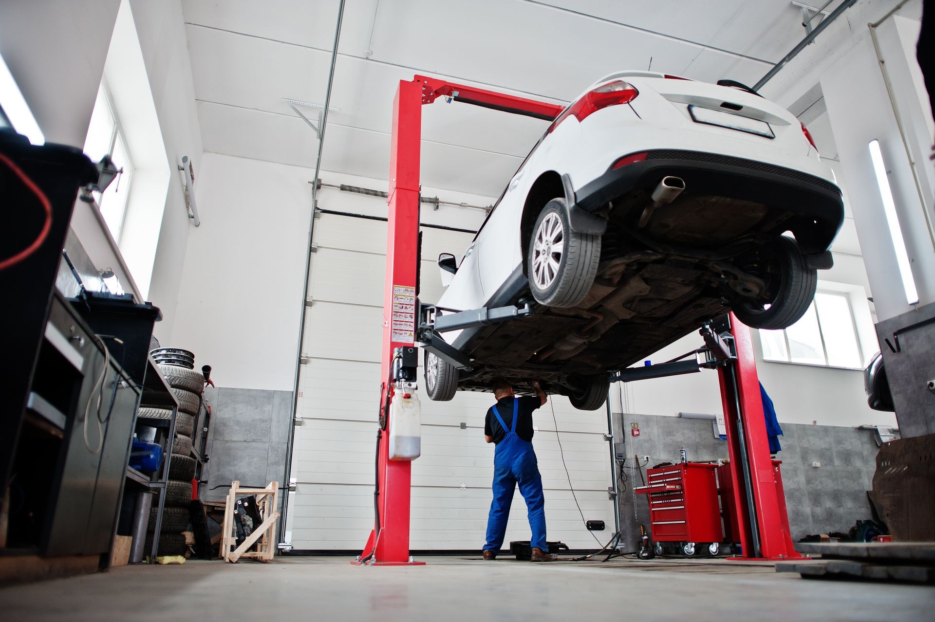 White car raised on a red hydraulic lift in an auto repair shop, with a mechanic working underneath.