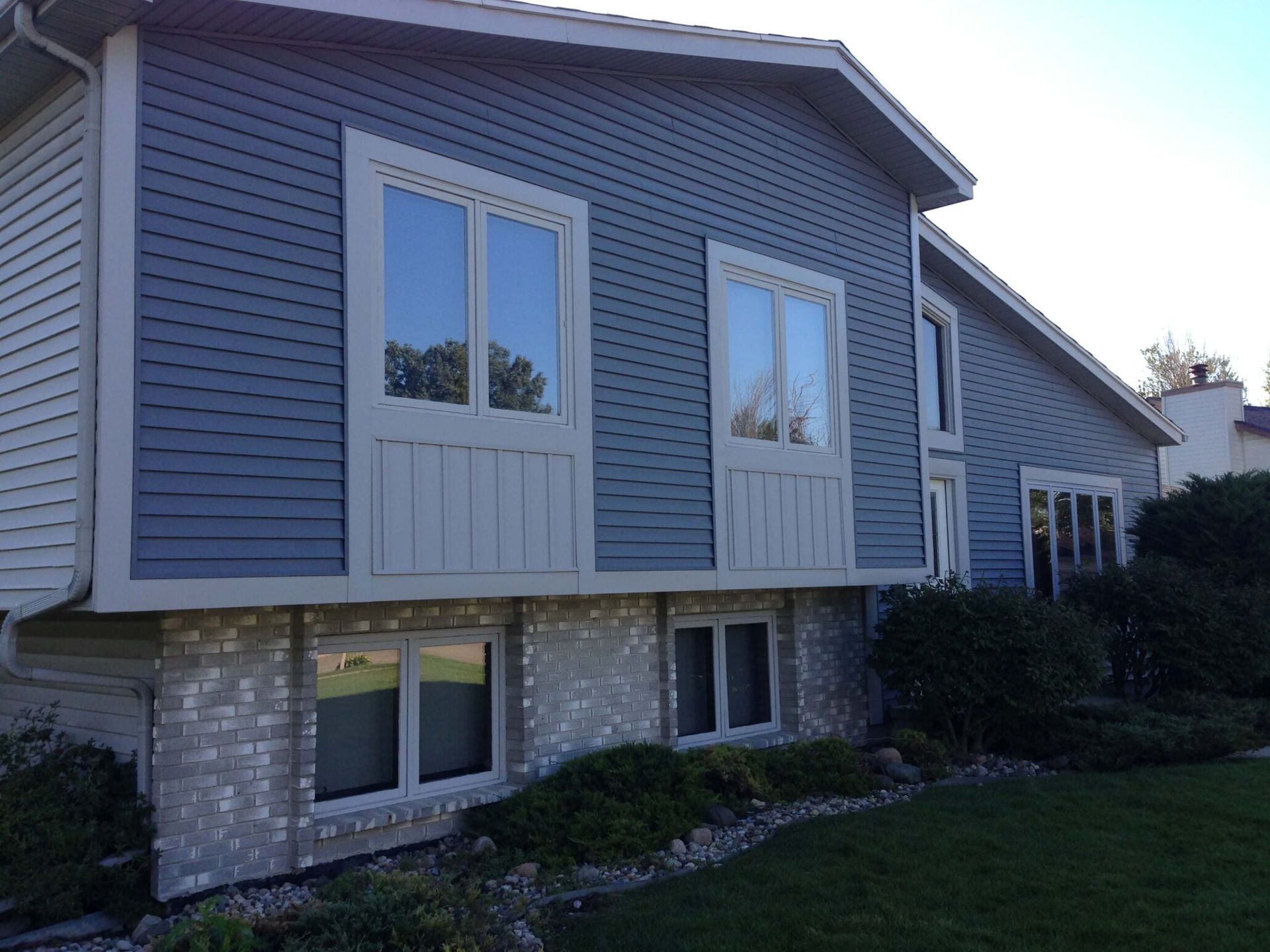 Gray house exterior with blue siding, white-trimmed windows, and stone foundation on a sloped lawn