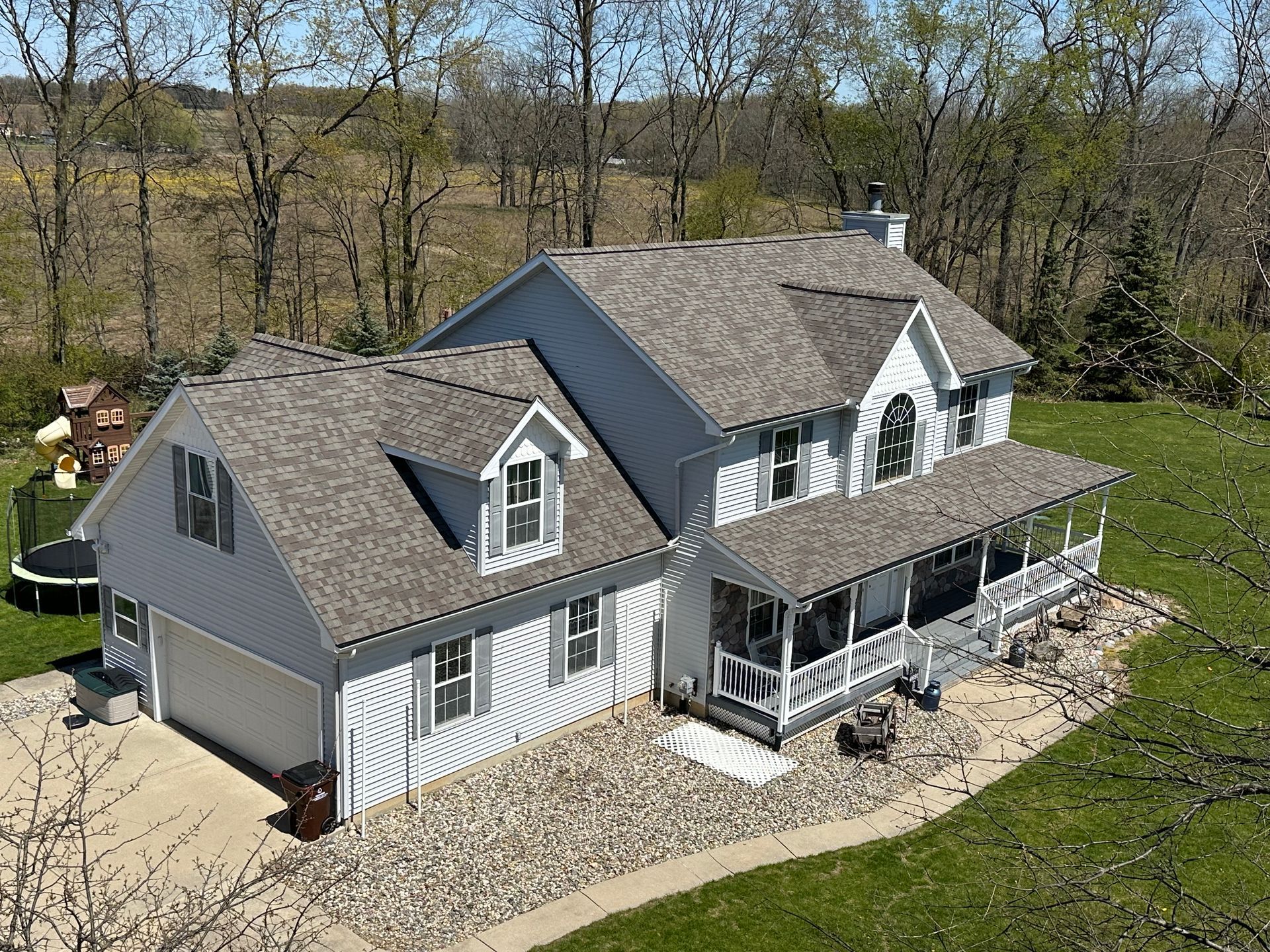 Gray two-story house with dormer windows, front porch, and stone landscaping in a wooded yard