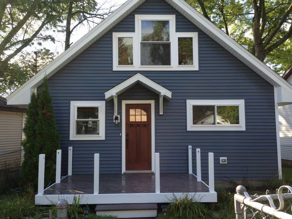 Blue house with front porch, brown door, and white trim on a tree-lined lot.