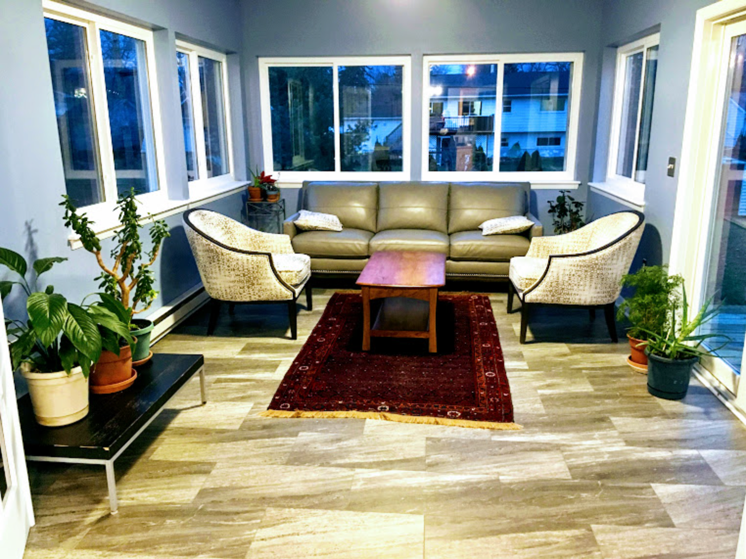 Sunroom with gray flooring, blue walls, a gray leather sofa, two patterned chairs, and many plants.