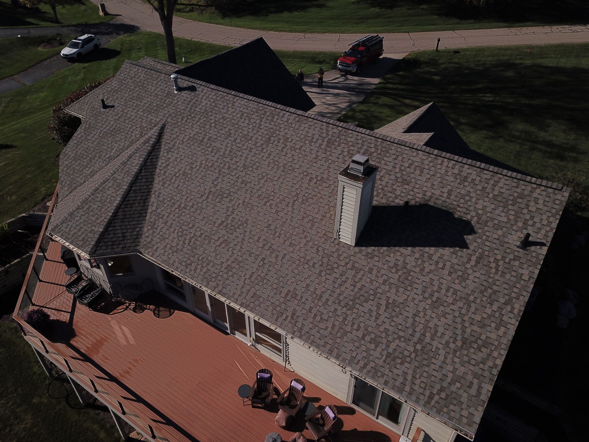 Aerial view of a house roof with a chimney and a patio below, surrounded by a grassy yard.