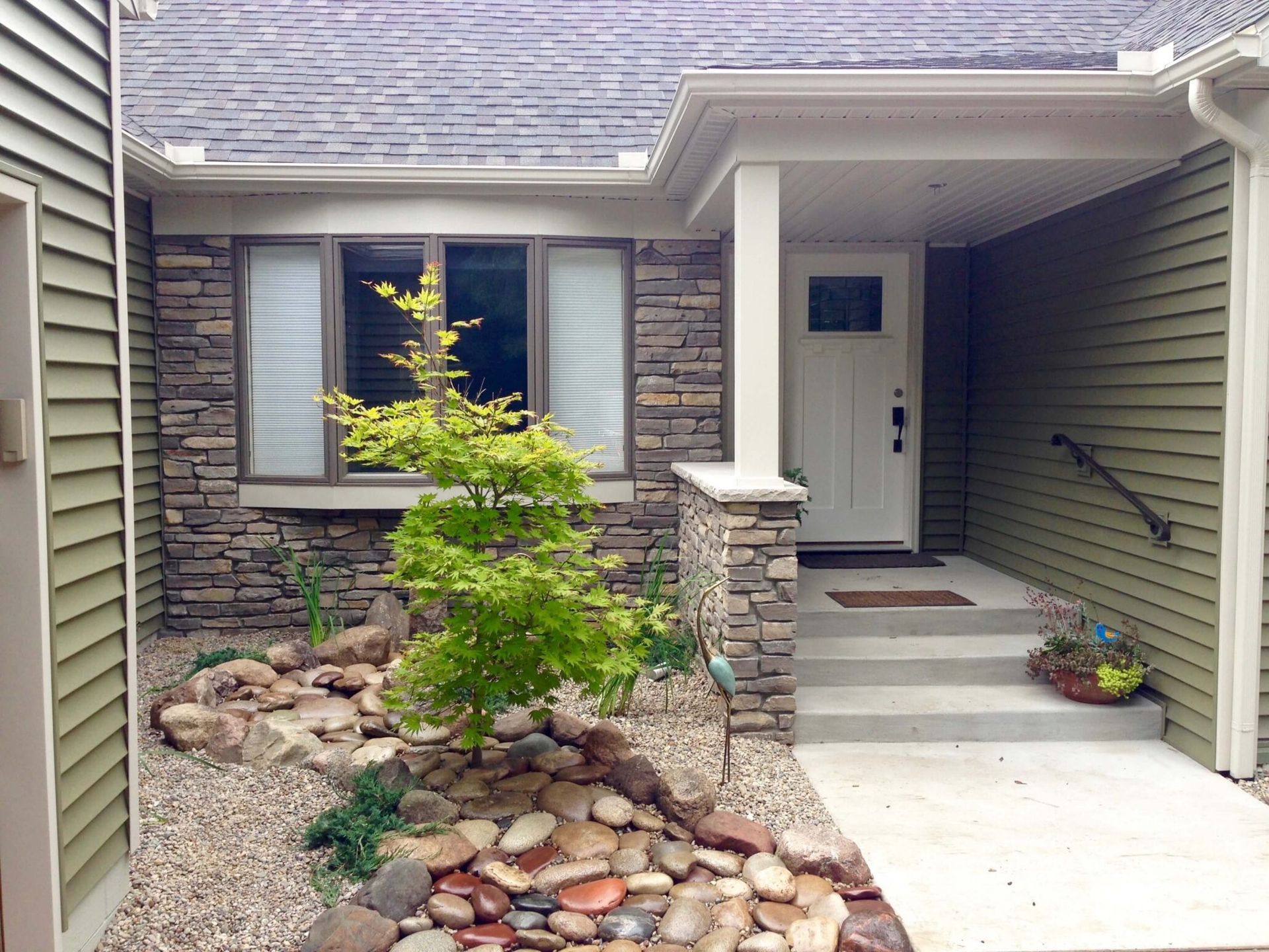 Front entry of a house with stone landscaping, concrete steps, and a white door under a covered porch